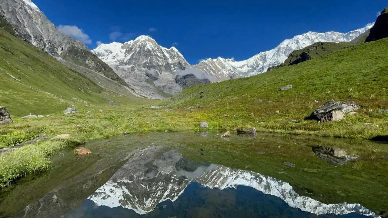 Lush green mountain valley with a clear pond reflecting snow-capped peaks under a bright sky