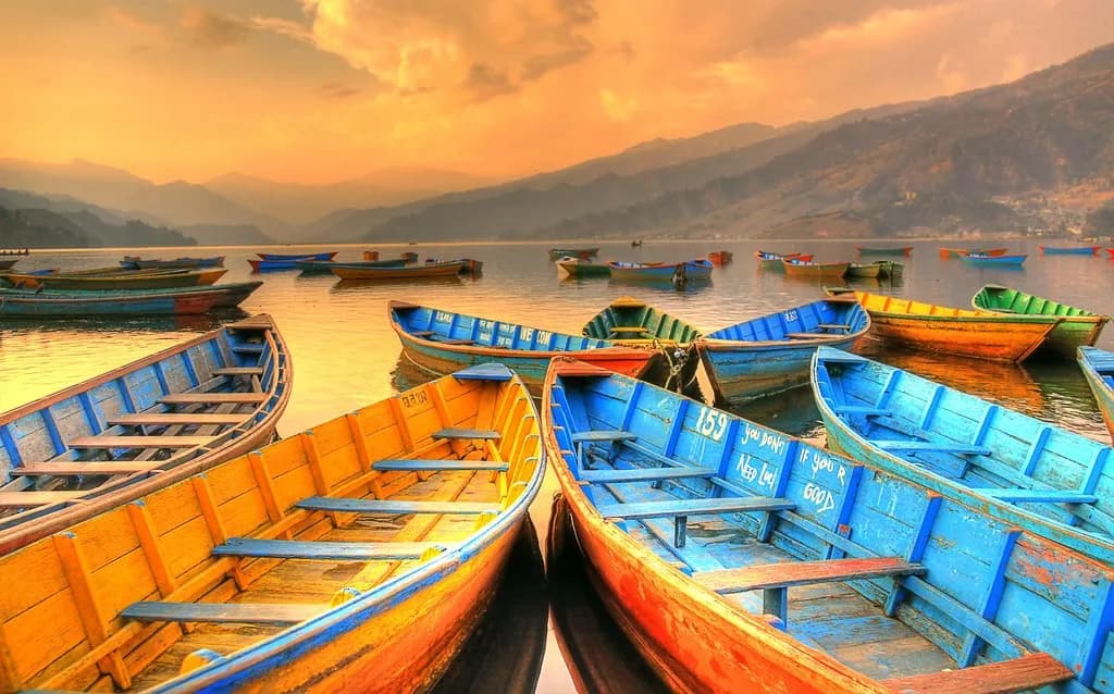 Vibrant yellow and blue wooden boats moored on Phewa Lake in Pokhara Nepal during a bright golden sunset with the Himalayan foothills in the distance