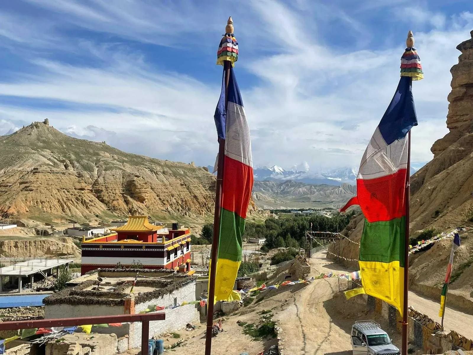 Tall vertical prayer flags overlooking a dusty road and a traditional village nestled in the valley